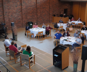 Overhead view of the chapel space with 3 people on the stage for a panel and the audience at round tables. 